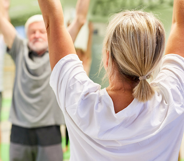 Eine Frau steht mit dem Rücken zum Fotografen und hebt die Arme beim Sport, im Hintergrund eine kleine Gruppe, die das nachmacht Eine Frau steht mit dem Rücken zum Fotografen und hebt die Arme beim Sport, im Hintergrund eine kleine Gruppe, die das nachmacht